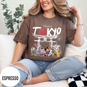 Puede incluir: Camiseta color espresso con un dise&ntilde;o de Disneyland Tokio, con personajes ic&oacute;nicos. El dise&ntilde;o incluye la palabra "TOKYO" en blanco, un c&iacute;rculo rojo y una puerta japonesa. La camiseta se lleva con vaqueros azules.