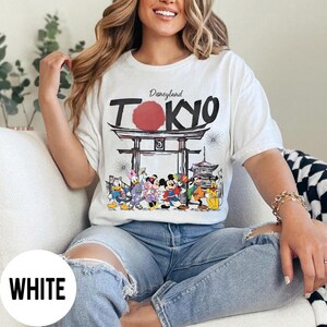 Puede incluir: Camiseta blanca con un dise&ntilde;o de Disneyland Tokio con Mickey Mouse y sus amigos. El dise&ntilde;o incluye un sol rojo, una puerta japonesa y fuegos artificiales. La camiseta es de estilo casual.