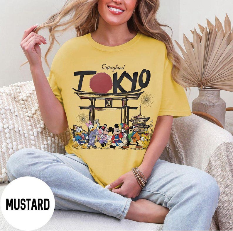 Puede incluir: Camiseta de color mostaza con el texto "Disneyland TOKYO" sobre un gr&aacute;fico de una puerta japonesa y personajes de dibujos animados. La camiseta tiene cuello redondo y mangas cortas. La camiseta la lleva una persona.