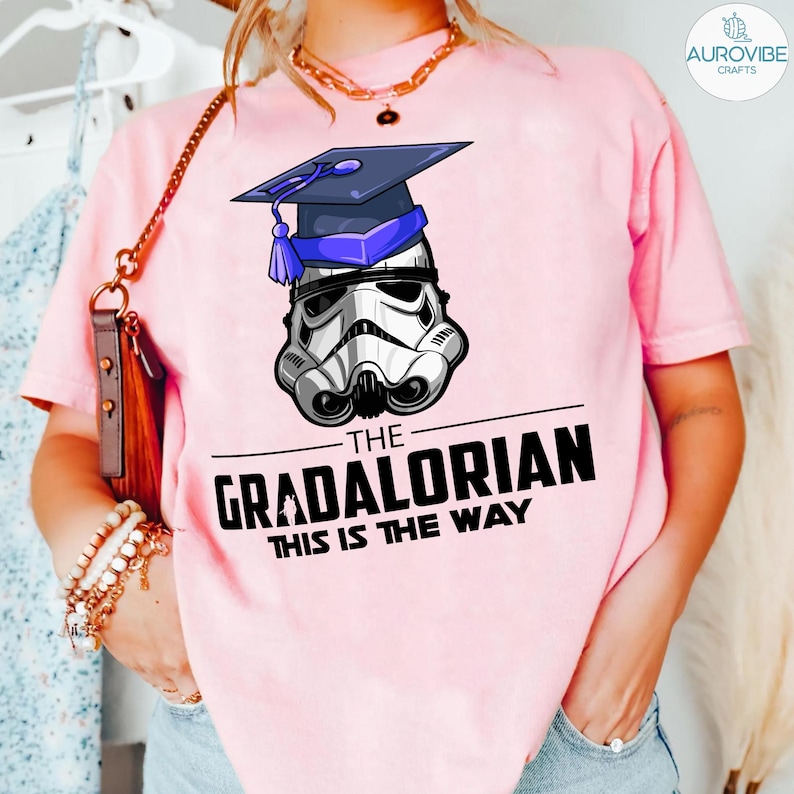 Pu&ograve; includere: T-shirt rosa chiaro con grafica di un elmetto da Stormtrooper con cappello di laurea. Il testo recita "THE GRADALORIAN THIS IS THE WAY" in nero. La maglietta &egrave; a girocollo.