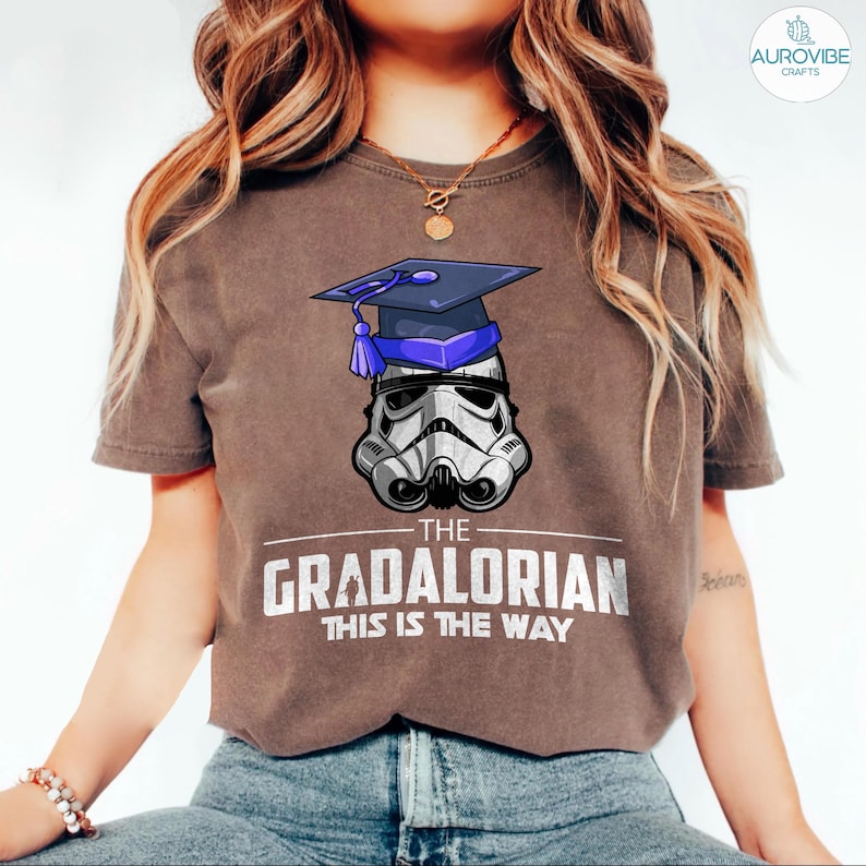 Pu&ograve; includere: T-shirt marrone con un casco da Stormtrooper con cappello di laurea. Il testo recita "The Gradalorian, This is the Way" in bianco. Un design divertente per i fan di Star Wars.
