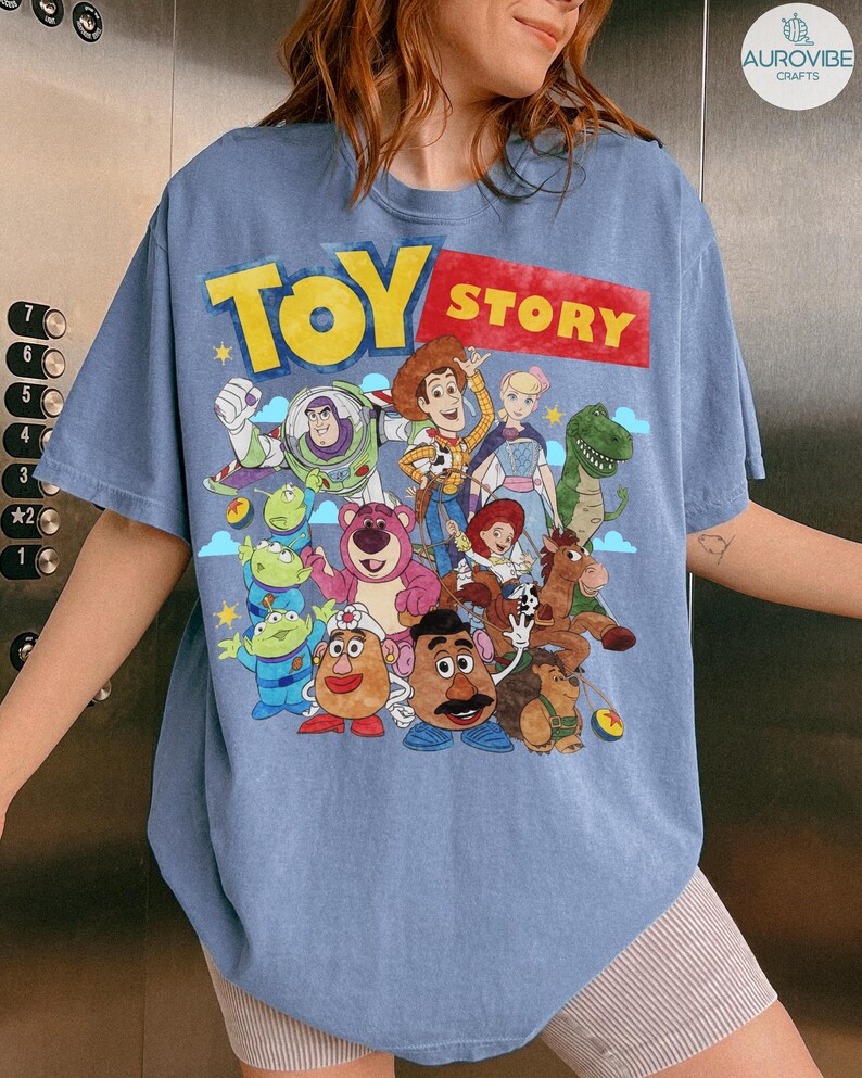 May include: A light blue t-shirt featuring the words "TOY STORY" in yellow and red, with a graphic of characters from the animated film. The shirt has a crew neck and short sleeves.