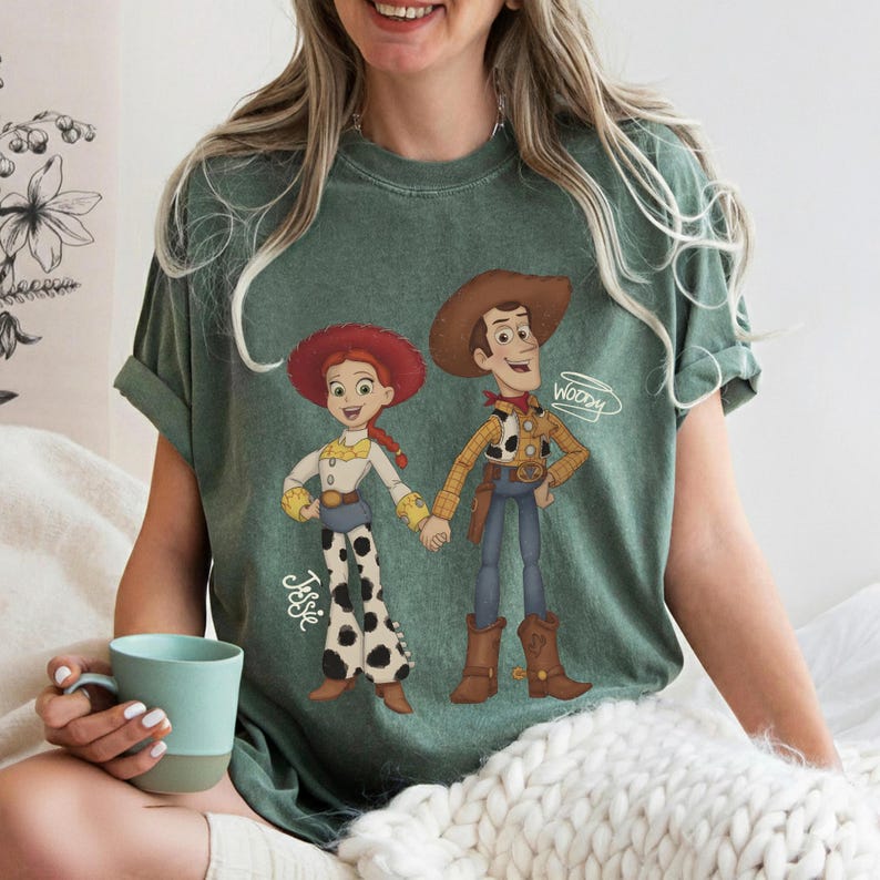 Może przedstawiać: Zielona koszulka z kresk&oacute;wkową ilustracją Jessie i Woody z Toy Story, trzymających się za ręce. Jessie ma na sobie czerwony kapelusz, białą koszulę i czarno-białe spodnie. Woody ma na sobie kapelusz kowbojski, ż&oacute;łtą koszulę i niebieskie spodnie. Na koszulce widnieją imiona "Jessie" i "Woody".