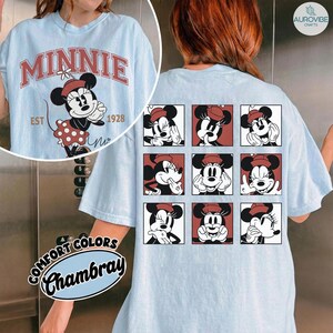 May include: Light blue t-shirt featuring a Minnie Mouse graphic. The front displays "MINNIE" in red lettering with a Minnie Mouse illustration and the text "EST 1928". The back has a grid of Minnie Mouse images. The shirt is made of Comfort Colors Chambray fabric.