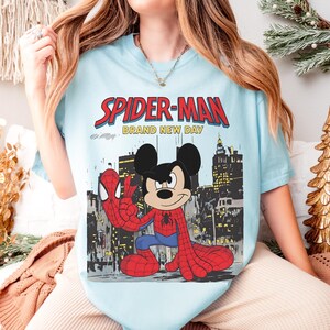 May include: Light blue t-shirt featuring a graphic of Mickey Mouse dressed as Spider-Man, with the words "SPIDER-MAN BRAND NEW DAY" in red and white. The design includes a cityscape background.