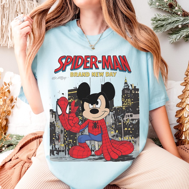 May include: Light blue t-shirt featuring a graphic of Mickey Mouse dressed as Spider-Man, with the words "SPIDER-MAN BRAND NEW DAY" in red and white. The design includes a cityscape background.