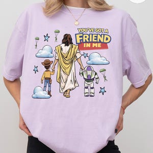 May include: Light purple t-shirt with the text "YOU'VE GOT A FRIEND IN ME". The graphic features cartoon illustrations of Jesus, a cowboy, and a space ranger, with clouds, stars, and parachutes.