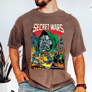 May include: Brown t-shirt featuring a comic book-style graphic with the text "SECRET WARS." The graphic depicts various superheroes and villains, including Doctor Doom, Spider-Man, and Captain America. The shirt has a classic fit.