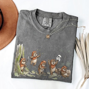 May include: Gray Comfort Colors t-shirt featuring a whimsical illustration of Ewoks in a forest setting. The design includes Ewoks with weapons and a Stormtrooper helmet. A silver necklace with a pendant is draped on the shirt.
