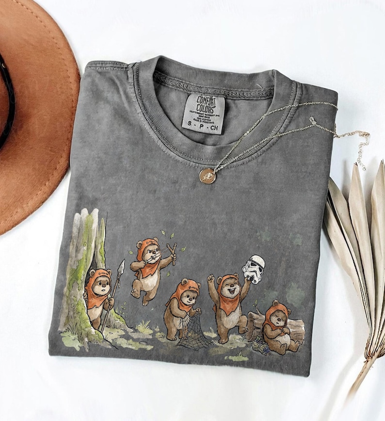 May include: Gray Comfort Colors t-shirt featuring a whimsical illustration of Ewoks in a forest setting. The design includes Ewoks with weapons and a Stormtrooper helmet. A silver necklace with a pendant is draped on the shirt.