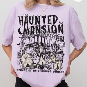 May include: A light purple t-shirt featuring a black graphic of The Haunted Mansion, with the text "Tomb Sweet Tomb" and "Beware of Hitchhiking Ghosts." The design includes illustrations of ghosts and bats.