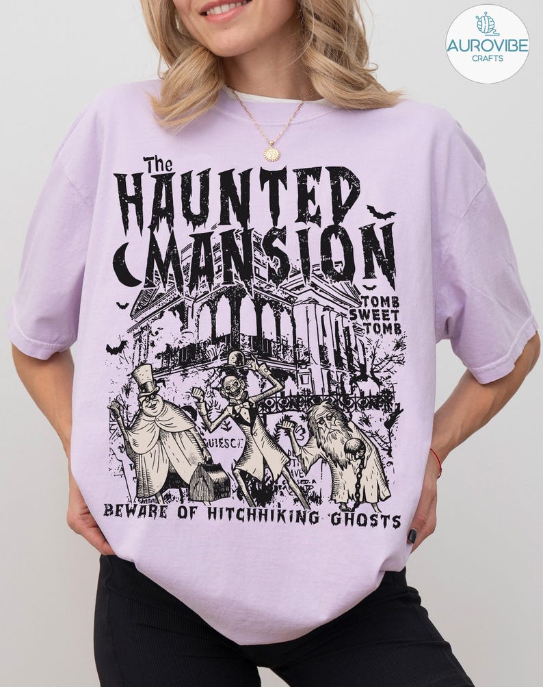 May include: A light purple t-shirt featuring a black graphic of The Haunted Mansion, with the text "Tomb Sweet Tomb" and "Beware of Hitchhiking Ghosts." The design includes illustrations of ghosts and bats.