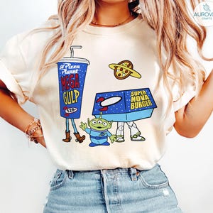 以下が含まれることがあります： クリーム色のTシャツ。青いメガガルフカップ、ピザプラネット、スーパーノヴァバーガーボックス、緑色のエイリアンが描かれた漫画のデザイン。レトロな宇宙をテーマにしたデザインです。