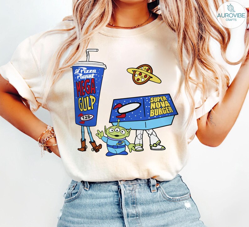 以下が含まれることがあります： クリーム色のTシャツ。青いメガガルフカップ、ピザプラネット、スーパーノヴァバーガーボックス、緑色のエイリアンが描かれた漫画のデザイン。レトロな宇宙をテーマにしたデザインです。