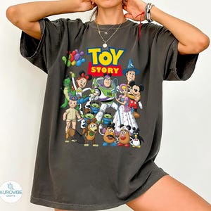 Op de afbeelding: Donkergrijs T-shirt met het "Toy Story" logo in geel en blauw. Het shirt toont een kleurrijke afbeelding van verschillende personages uit de animatiefilm, waaronder Buzz Lightyear, Woody en Mickey Mouse.