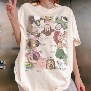 K&ouml;nnte beinhalten: Cremefarbenes T-Shirt mit einer bunten Grafik von Charakteren aus dem Animationsfilm "Toy Story". Das Design umfasst Woody, Buzz Lightyear, Jessie, Rex und andere Charaktere mit ihren Namen darunter. Das Shirt hat kurze &Auml;rmel.