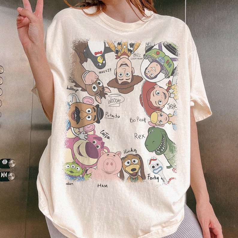 K&ouml;nnte beinhalten: Cremefarbenes T-Shirt mit einer bunten Grafik von Charakteren aus dem Animationsfilm "Toy Story". Das Design umfasst Woody, Buzz Lightyear, Jessie, Rex und andere Charaktere mit ihren Namen darunter. Das Shirt hat kurze &Auml;rmel.