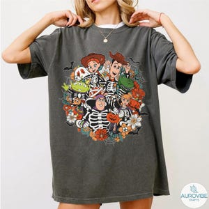 May include: A charcoal gray short-sleeve t-shirt featuring a graphic print of various animated characters wearing skeleton costumes. The design includes orange flowers, spider webs, and bats, creating a festive seasonal arrangement on the front.
