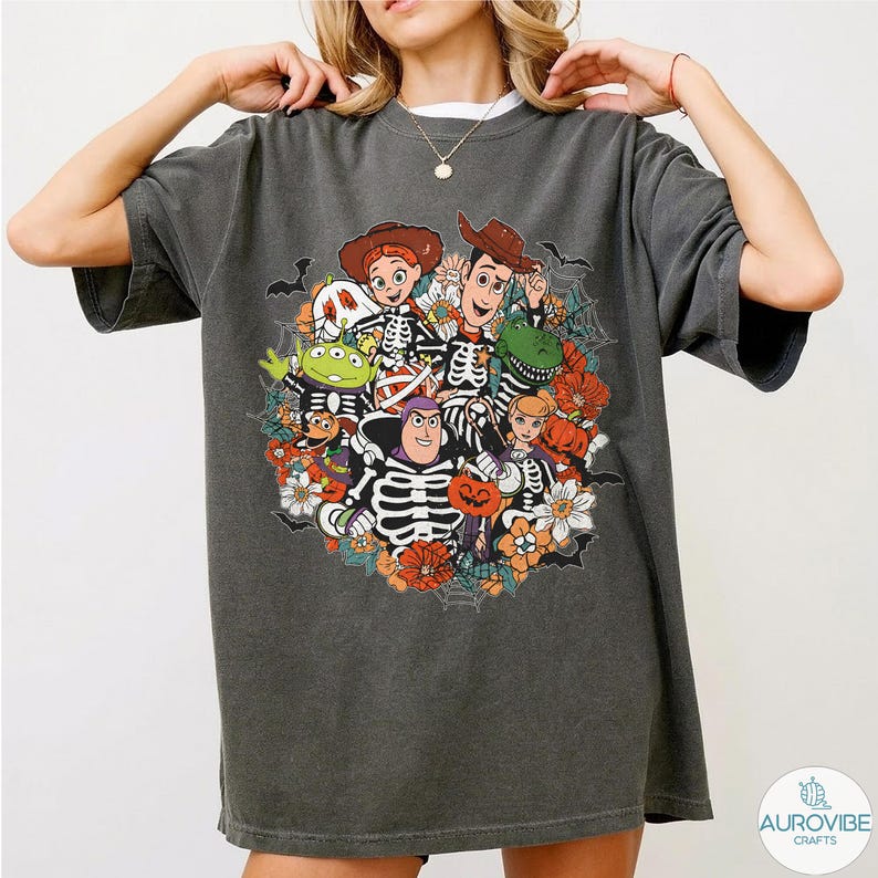 May include: A charcoal gray short-sleeve t-shirt featuring a graphic print of various animated characters wearing skeleton costumes. The design includes orange flowers, spider webs, and bats, creating a festive seasonal arrangement on the front.