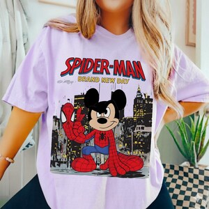 May include: A light purple t-shirt featuring a graphic of Mickey Mouse dressed as Spider-Man. The design includes the words "SPIDER-MAN" in red and "BRAND NEW DAY" in white, set against a cityscape backdrop.