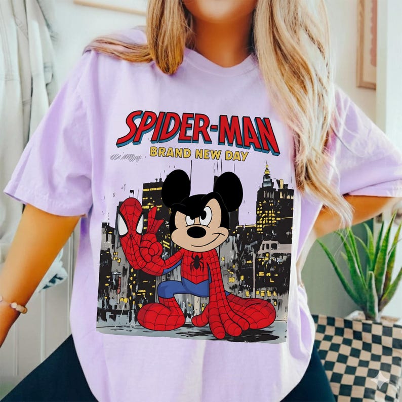 May include: A light purple t-shirt featuring a graphic of Mickey Mouse dressed as Spider-Man. The design includes the words "SPIDER-MAN" in red and "BRAND NEW DAY" in white, set against a cityscape backdrop.