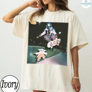 Puede incluir: Camiseta color marfil con un gr&aacute;fico de un skater y Grogu patinando en un skate park. El skater lleva casco y armadura. Grogu lleva un abrigo color beige. La camiseta tiene un corte holgado y la palabra "Ivory" est&aacute; impresa en la parte inferior.