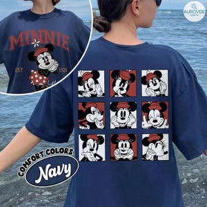 May include: Navy blue t-shirt featuring Minnie Mouse graphics. The front displays "MINNIE" with a vintage design and the year "1928". The back showcases a grid of Minnie Mouse expressions. The shirt has a "COMFORT COLORS" label.