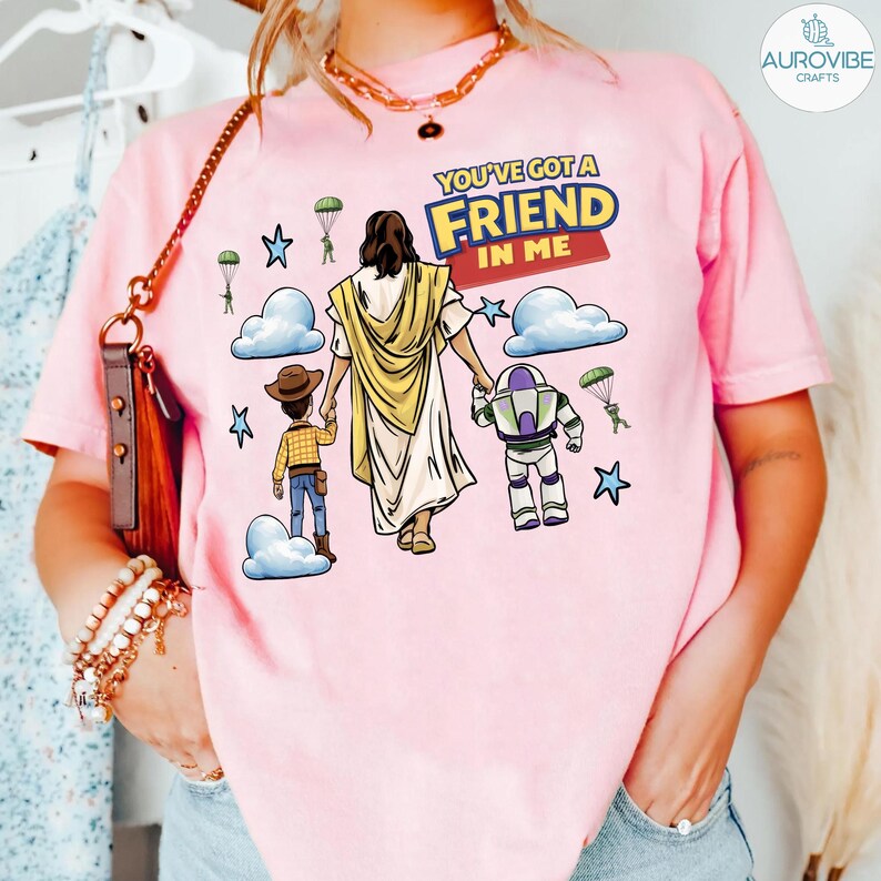 May include: Pink t-shirt featuring a graphic of Jesus holding hands with cartoon characters, including a cowboy and a space ranger. The text "YOU'VE GOT A FRIEND IN ME" is displayed above the characters. The shirt has a crew neck and short sleeves.