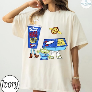 以下が含まれることがあります： アイボリー色のTシャツ。青いメガガルのプカップ、ピザプラネット、エイリアン、スーパーノヴァバーガーの箱が描かれた漫画のデザイン。デザインは遊び心があり、アニメーションスタイルです。