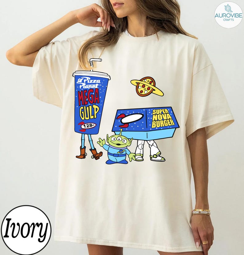 以下が含まれることがあります： アイボリー色のTシャツ。青いメガガルのプカップ、ピザプラネット、エイリアン、スーパーノヴァバーガーの箱が描かれた漫画のデザイン。デザインは遊び心があり、アニメーションスタイルです。