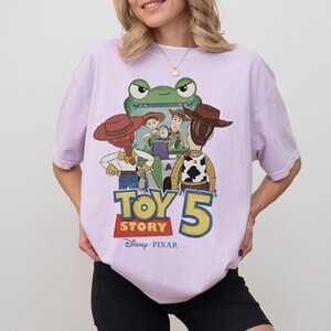 K&ouml;nnte beinhalten: Helllila T-Shirt mit einem Toy Story 5-Motiv. Das Design zeigt Charaktere wie Woody, Buzz Lightyear und Jessie, mit dem Filmtitel in einer kr&auml;ftigen, farbenfrohen Schrift. Das Disney Pixar-Logo ist ebenfalls zu sehen.