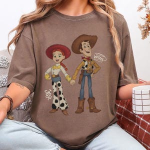 Może przedstawiać: Brązowa koszulka z kresk&oacute;wkową ilustracją Jessie i Woody z Toy Story. Jessie ma na sobie czerwony kapelusz, białą koszulę i spodnie w krowie łaty. Woody nosi kapelusz kowbojski, ż&oacute;łtą koszulę i niebieskie dżinsy. Na koszulce widnieją imiona "Jessie" i "Woody".