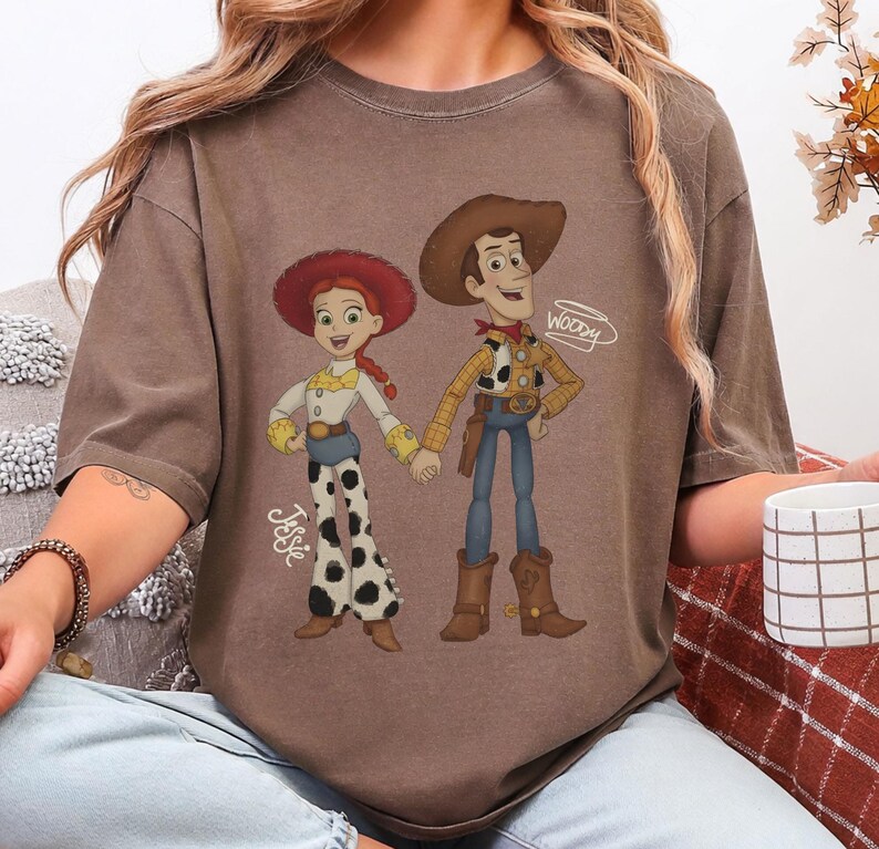 Może przedstawiać: Brązowa koszulka z kresk&oacute;wkową ilustracją Jessie i Woody z Toy Story. Jessie ma na sobie czerwony kapelusz, białą koszulę i spodnie w krowie łaty. Woody nosi kapelusz kowbojski, ż&oacute;łtą koszulę i niebieskie dżinsy. Na koszulce widnieją imiona "Jessie" i "Woody".