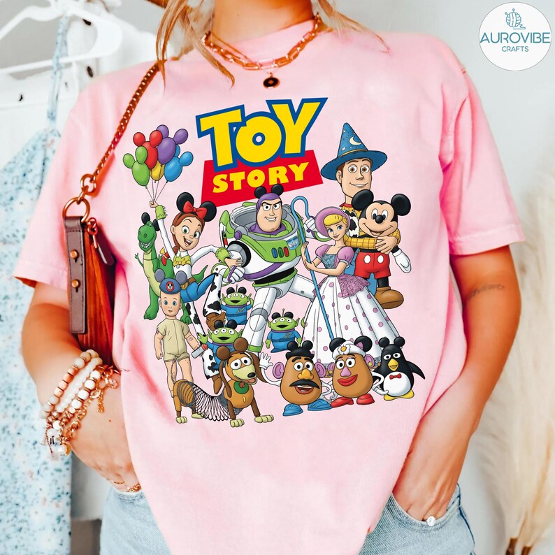 Op de afbeelding: Zachtroze T-shirt met een kleurrijke afbeelding van personages uit de film "Toy Story". Het ontwerp bevat het logo van de film en personages zoals Buzz Lightyear, Woody en Mickey Mouse. Het T-shirt heeft een ronde hals.