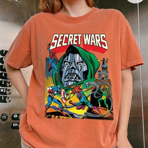 May include: Orange t-shirt featuring a comic book graphic with the text "SECRET WARS." The graphic depicts various superheroes in action, including Spider-Man and Captain America, with a large metal-masked figure in the background.