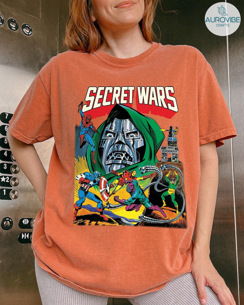 May include: Orange t-shirt featuring a comic book graphic with the text "SECRET WARS." The graphic depicts various superheroes in action, including Spider-Man and Captain America, with a large metal-masked figure in the background.