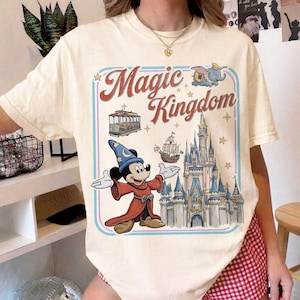 May include: Cream-colored t-shirt featuring a colorful graphic with the words "Magic Kingdom." The design includes Mickey Mouse in a wizard outfit, a castle, a trolley car, a ship, and Dumbo the elephant. The design is framed with a blue and pink border.