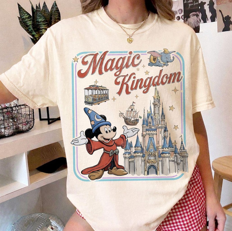May include: Cream-colored t-shirt featuring a colorful graphic with the words "Magic Kingdom." The design includes Mickey Mouse in a wizard outfit, a castle, a trolley car, a ship, and Dumbo the elephant. The design is framed with a blue and pink border.