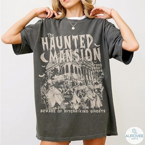 May include: Dark gray t-shirt featuring a graphic of The Haunted Mansion, with the text "The Haunted Mansion Tomb Sweet Tomb" and "Beware of Hitchhiking Ghosts." The design includes a spooky house, bats, and ghostly figures.