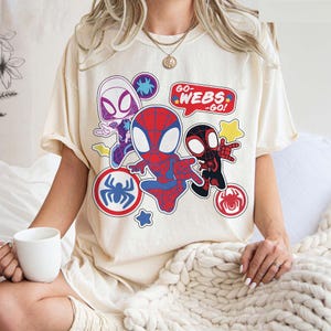 Puede incluir: Camiseta color crema con un gr&aacute;fico colorido de personajes de Spider-Man. El dise&ntilde;o incluye a Spider-Gwen, Spider-Man y Miles Morales, con el texto "GO WEBS - GO!". La camiseta tiene mangas cortas.