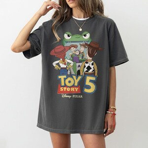 K&ouml;nnte beinhalten: Dunkelgraues T-Shirt mit einem farbenfrohen Toy Story 5-Motiv. Das Design zeigt Charaktere wie Woody und Buzz Lightyear sowie den Filmtitel und das Disney Pixar-Logo. Das T-Shirt hat eine lockere Passform.