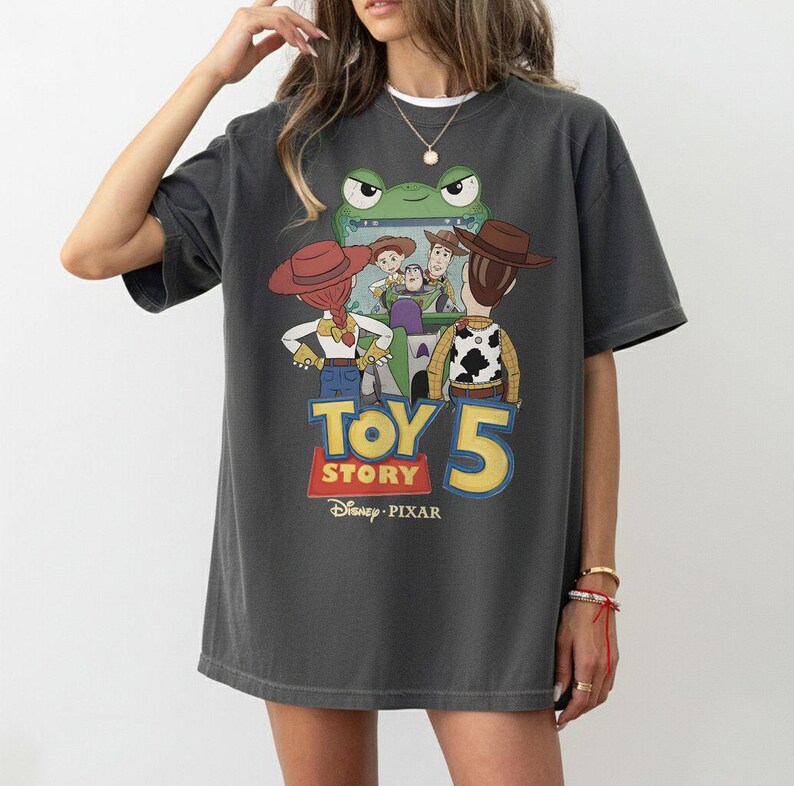 K&ouml;nnte beinhalten: Dunkelgraues T-Shirt mit einem farbenfrohen Toy Story 5-Motiv. Das Design zeigt Charaktere wie Woody und Buzz Lightyear sowie den Filmtitel und das Disney Pixar-Logo. Das T-Shirt hat eine lockere Passform.