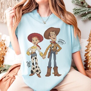Może przedstawiać: Jasnoniebieska koszulka z kresk&oacute;wkową ilustracją Woody'ego i Jessie z Toy Story, trzymających się za ręce. Jessie ma na sobie białą koszulę, spodnie w krowie łaty i czerwony kapelusz. Woody ma na sobie ż&oacute;łtą koszulę, niebieskie dżinsy i brązowy kapelusz. Na koszulce widnieje napis "Woody".