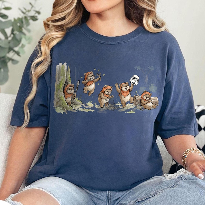 May include: Navy blue t-shirt featuring a whimsical illustration of Ewoks in a forest setting. The design includes several Ewoks, some holding sticks and a Stormtrooper helmet. The artwork is detailed with trees, foliage, and a log.