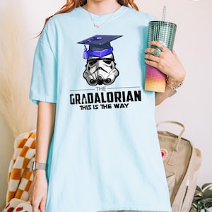 Pu&ograve; includere: T-shirt azzurra con un casco da Stormtrooper con cappello di laurea. Il testo "THE GRADALORIAN THIS IS THE WAY" &egrave; stampato sotto. La maglietta &egrave; in stile casual.