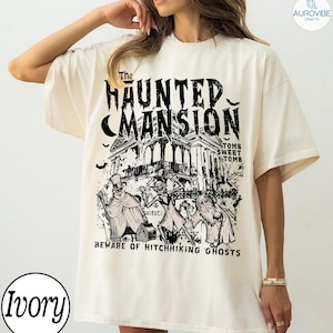 May include: Ivory-colored t-shirt featuring a black graphic of a haunted mansion with the text "The Haunted Mansion". Below the mansion are illustrations of ghosts and the phrase "Beware of Hitchhiking Ghosts".