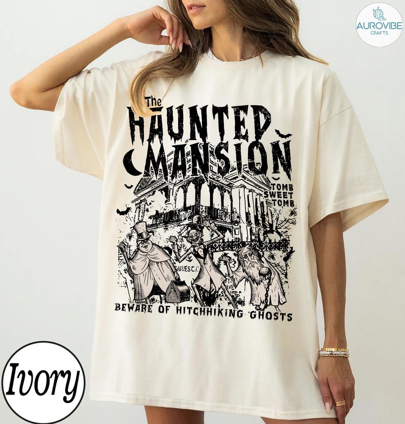 May include: Ivory-colored t-shirt featuring a black graphic of a haunted mansion with the text "The Haunted Mansion". Below the mansion are illustrations of ghosts and the phrase "Beware of Hitchhiking Ghosts".
