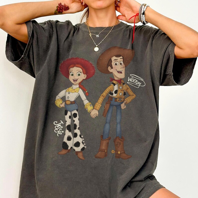 Może przedstawiać: Ciemnoszara koszulka z kresk&oacute;wkową ilustracją Jessie i Woody'ego z Toy Story. Jessie ma na sobie czerwony kapelusz, białą koszulę i spodnie w krowie łaty. Woody ma na sobie kapelusz kowbojski, ż&oacute;łtą koszulę i niebieskie dżinsy. Na koszulce widnieje napis "Jessie".