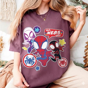 Puede incluir: Camiseta granate con ilustraciones de dibujos animados de personajes de Spider-Man. El dise&ntilde;o incluye a Spider-Man, Spider-Gwen y Miles Morales, con bocadillos que dicen "GO-WEBS-GO!" y emblemas de ara&ntilde;a.