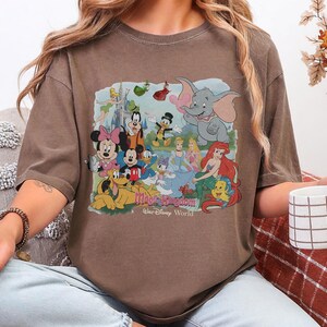 Puede incluir: Camiseta marr&oacute;n de manga corta con un gr&aacute;fico colorido de personajes de Disney, incluyendo Mickey y Minnie Mouse, Donald Duck y Dumbo. El gr&aacute;fico incluye el texto "Magic Kingdom" y "Walt Disney World".